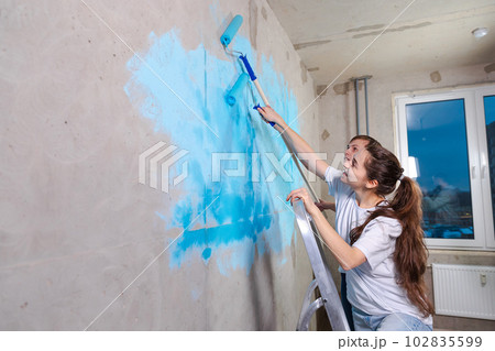 Couple in new home during repair works painting wall together. Happy family holding paint roller painting wall with blue color paint in new house. Home renovation DIY renew home concept Couple in new home during repair works painting wall together. Happy family holding paint roller painting wall with blue color paint in new house. Home renovation DIY renew home concept 102835599