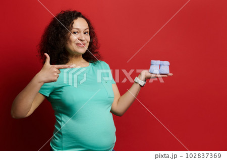 Charming pregnant woman points aside cute baby booties on her hand, smiles looking at camera, isolated on red background. Happy expectant mother expressing positive emotions of pregnancy and maternity 102837369