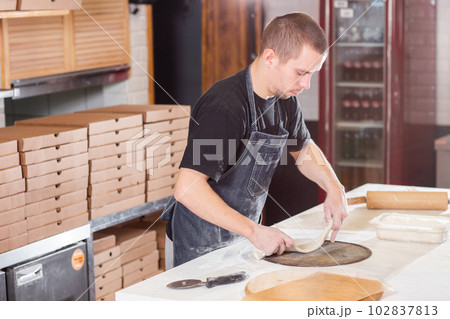 The chef prepares pizza. Raw pizza ready to bake. Cook in a blue apron in the kitchen. with a shovel in his hands. boxes for food delivery on background. The chef prepares pizza. Raw pizza ready to bake. Cook in a blue apron in the kitchen. with a shovel in his hands. boxes for food delivery on background. 102837813