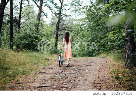 Walk in the woods for wellness, Health benefits of forests. Immerse Yourself in a Forest. A young girl rolls a bicycle along a forest path in tree wood Walk in the woods for wellness, Health benefits of forests. Immerse Yourself in a Forest. A young girl rolls a bicycle along a forest path in tree wood 102837816