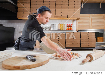 The chef prepares pizza. Raw pizza ready to bake. Cook in a blue apron in the kitchen. with a shovel in his hands. boxes for food delivery on background. The chef prepares pizza. Raw pizza ready to bake. Cook in a blue apron in the kitchen. with a shovel in his hands. boxes for food delivery on background. 102837844