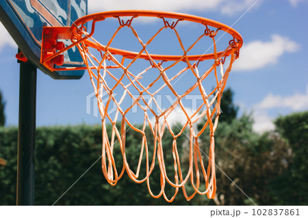 Basketball hoop on backyard against blue sky. Sports recreation and healthy living in nature. Orange red rope net on a street. Equipment on a playground. Training tools on stadium. Play outdoors. Basketball hoop on backyard against blue sky. Sports recreation and healthy living in nature. Orange red rope net on a street. Equipment on a playground. Training tools on stadium. Play outdoors. 102837861