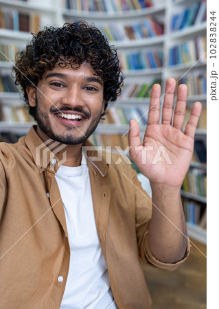 A young happy Spanish male student is talking on a video call from a phone in the library. Greets and waves at the camera, participates in the international student exchange program. Vertical photo. 102838244