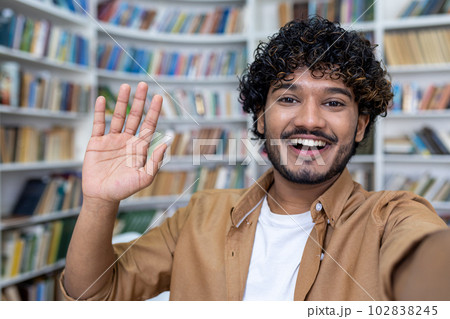 Student video call, hispanic man smiling and looking into phone camera, man using app for online communication with friends, student taking selfie photo in library. 102838245