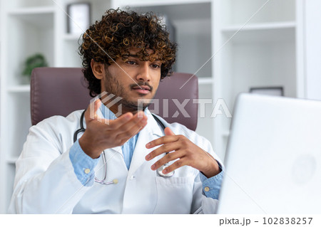 Close-up photo. A young hispanic doctor in a white coat and with a stethoscope consults patients online via video call from a laptop in the office. Talks, helps, supports. 102838257