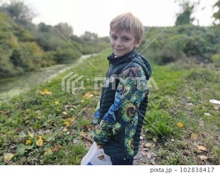 A blond boy, 8 years old, laughs in the fall on a walk by a swampy pond. A violent emotion of fun on the face of a child. Everyday warm clothes. Green jacket. 102838417