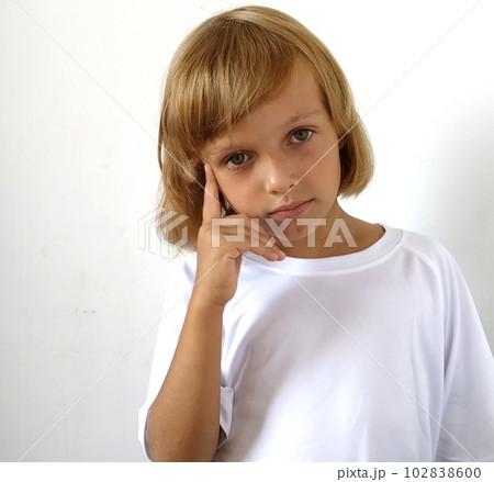 Little cute girl 6-7 years old with blond hair on a white background. Tanned skin. White T-shirt. Copy space. The girl raised her right hand and put her index finger to her temple. Hairstyle Bob. 102838600
