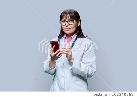 Mature female doctor in white lab coat using phone, on grey background 102840369