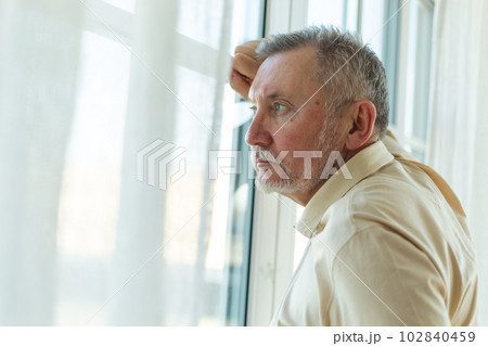 Sad tired lonely disappointed older senior man at home alone. Unhappy grandfather looking sadly outside window experiencing grief relative death bad news. Stressed elder man suffering from loneliness 102840459