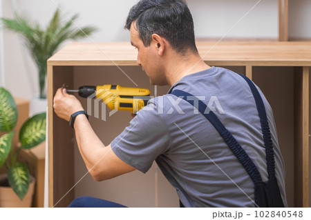 Wooden furniture industry man attaching door hinge to wooden cabinet side with screwdriver firm worker in overalls assembling new furniture for living room interior 102840548