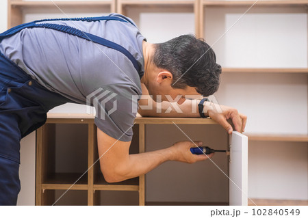 Furniture fixing services professional worker in uniform screwing into panel made of quality wood material man uses tools sitting on laminate floor in room 102840549