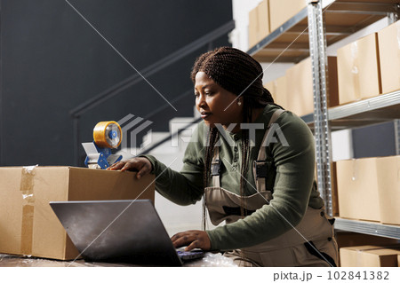 Stockroom supervisor preparing customers orders using cardboard boxes, checking shipping details on laptop computer in storehouse. African american employee working at products inventory report 102841382