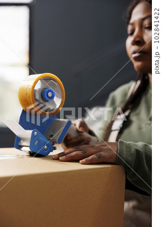 African american worker preparing customers packages for delivery using adhesive tape, doing products quality control in storehouse. Stockroom employee working at online orders. Close up 102841422
