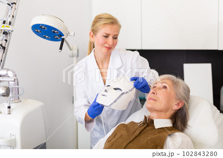 Cosmetologist performing anti-aging face phototherapy procedure for aged woman Cosmetologist performing anti-aging face phototherapy procedure for aged woman 102843280