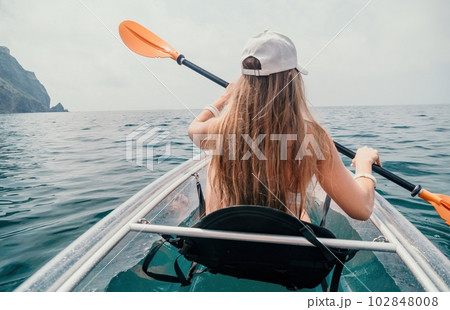 Woman in kayak back view. Happy young woman with long hair floating in transparent kayak on the crystal clear sea. Summer holiday vacation and cheerful female people relaxing having fun on the boat Woman in kayak back view. Happy young woman with long hair floating in transparent kayak on the crystal clear sea. Summer holiday vacation and cheerful female people relaxing having fun on the boat 102848008