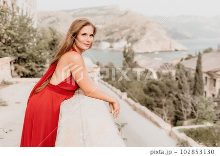 Woman travel portrait. Happy woman with long hair looking at camera and smiling. Close up portrait cute woman in a res long dress posing on backdrop of old travel city near sea 102853130