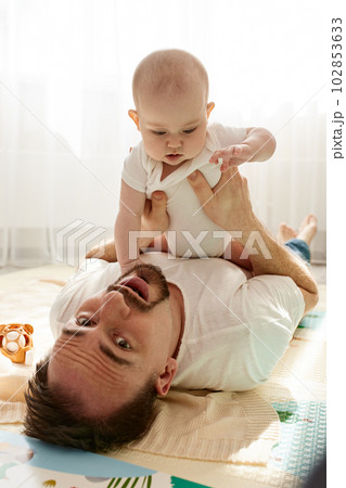 Happy dad holds his newborn baby in his arms while lying on the floor in a bright child's room. The father looks at the child with a loving look. Happy childhood and fatherhood. 102853633