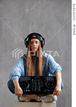 Studio portrait of a young pretty long-haired DJ girl in a blue sweater, loose jeans and a black baseball cap posing while sitting with a DJ console in her hands. Music, fun and creativity. Studio portrait of a young pretty long-haired DJ girl in a blue sweater, loose jeans and a black baseball cap posing while sitting with a DJ console in her hands. Music, fun and creativity. 102853678