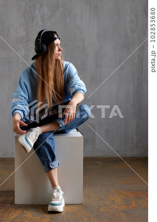 Studio portrait of a young pretty long-haired DJ girl in a blue sweater, loose jeans and a black baseball cap posing while sitting with a DJ console in her hands. Music, fun and creativity. Studio portrait of a young pretty long-haired DJ girl in a blue sweater, loose jeans and a black baseball cap posing while sitting with a DJ console in her hands. Music, fun and creativity. 102853680