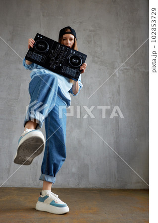 A young pretty long haired hip hop girl in a blue sweater, loose jeans and a black baseball cap holds a dj console in her hands and dances vigorously. Music, fun and creativity. A young pretty long haired hip hop girl in a blue sweater, loose jeans and a black baseball cap holds a dj console in her hands and dances vigorously. Music, fun and creativity. 102853729