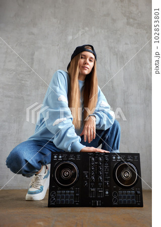 A young pretty long haired hip hop girl in a blue sweater, loose jeans and a black baseball cap is squatting and holding a dj console. Music, fun and creativity. 102853801