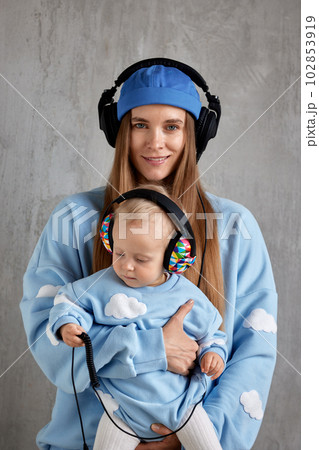 Portrait of a happy young mother and baby infant in her arms. Mom hugs her cute baby. Baby and mother in blue clothes and DJ headphones on their heads. 102853919
