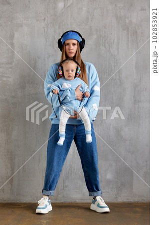 Full length portrait of a happy young mother and baby infant in her arms. Mom hugs her cute baby. Baby and mother in blue clothes and DJ headphones on their heads. 102853921
