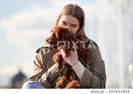A young smiling blonde girl in a stylish beige raincoat holds and hugs her pet toy poodle in her arms. The girl and the dog are resting in the park. Showing affection concept. 102853958