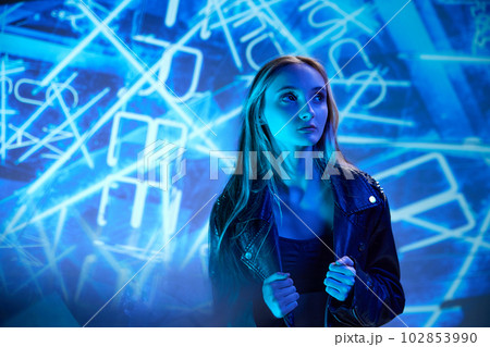 Portrait of young beautiful girl in leather jacket posing, looking away over blue background with abstract neon elements Portrait of young beautiful girl in leather jacket posing, looking away over blue background with abstract neon elements 102853990