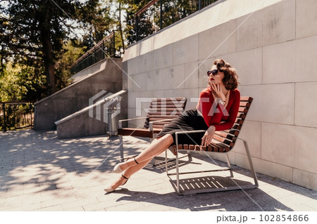 Portrait of a woman on the street. An attractive woman in glasses, a red blouse and a black skirt is sitting on a bench outside. Portrait of a woman on the street. An attractive woman in glasses, a red blouse and a black skirt is sitting on a bench outside. 102854866