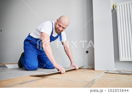 Male construction worker laying laminate wooden plank on floor underlayment. Man in work overalls kneeling down by the wall and installing laminate flooring in living room. Home renovation concept. Male construction worker laying laminate wooden plank on floor underlayment. Man in work overalls kneeling down by the wall and installing laminate flooring in living room. Home renovation concept. 102855644