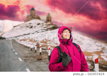 Stepantsminda, Georgia. Man Tourist Backpacker Traveler Photographer In Pink Jacket Posing Near Holy Trinity Church - Tsminda Sameba. Amazing Georgian Landmark In Early Winter. Bold Bright Sky Stepantsminda, Georgia. Man Tourist Backpacker Traveler Photographer In Pink Jacket Posing Near Holy Trinity Church - Tsminda Sameba. Amazing Georgian Landmark In Early Winter. Bold Bright Sky 102855929