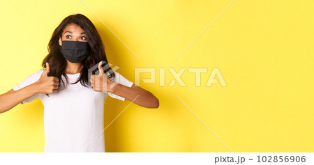 Concept of covid-19, social distancing and lifestyle. Image of cheerful african-american girl in black face mask, showing thumbs-up and looking happy at upper left corner, yellow background Concept of covid-19, social distancing and lifestyle. Image of cheerful african-american girl in black face mask, showing thumbs-up and looking happy at upper left corner, yellow background 102856906