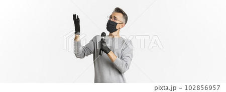 Concept of coronavirus, lifestyle and quarantine. Portrait of charismatic middle-aged man in medical mask and gloves, singing serenade in microphone, standing over white background 102856957