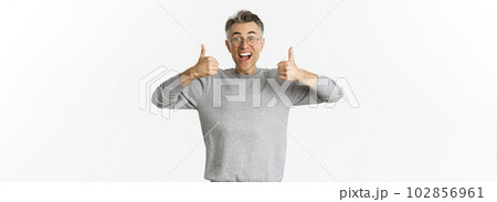 Portrait of handsome middle-aged man in glasses and grey sweater, looking excited and smiling, showing thumbs-up to approve and agree with something, like idea, standing over white background Portrait of handsome middle-aged man in glasses and grey sweater, looking excited and smiling, showing thumbs-up to approve and agree with something, like idea, standing over white background 102856961