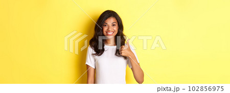 Portrait of happy and cute african-american girl, smiling pleased and showing thumbs-up in approval, like and approve something, standing over yellow background Portrait of happy and cute african-american girl, smiling pleased and showing thumbs-up in approval, like and approve something, standing over yellow background 102856975