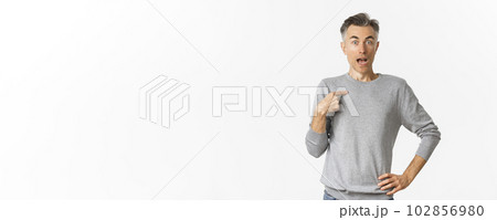 Portrait of handsome middle-aged gay man looking surprised, pointing at himself, being chosen, standing over white background in grey basic sweater Portrait of handsome middle-aged gay man looking surprised, pointing at himself, being chosen, standing over white background in grey basic sweater 102856980