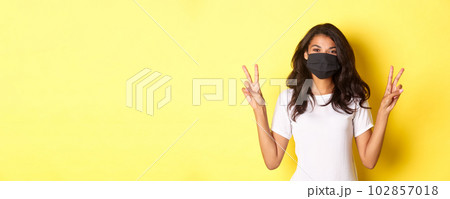 Concept of covid-19, social distancing and lifestyle. Portrait of beautiful african-american girl in black face mask, showing peace signs and smiling, standing over yellow background 102857018