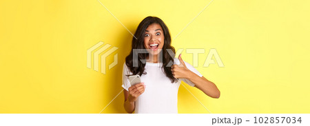 Portrait of amazed african-american girl, showing thumbs-up and using mobile phone, praising cool app, standing over yellow background Portrait of amazed african-american girl, showing thumbs-up and using mobile phone, praising cool app, standing over yellow background 102857034