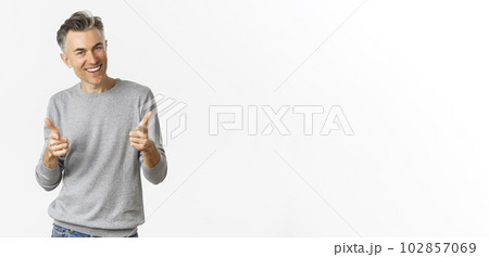 Cheeky middle-aged man with grey short hairstyle, pointing fingers at camera and smiling, praising good work or congratulating, standing over white background 102857069