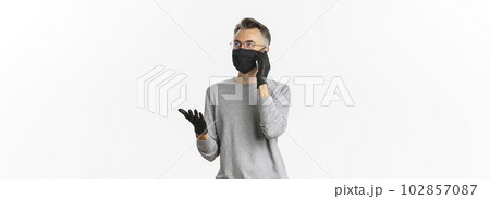 Concept of covid-19, social distancing and lifestyle. Image of handsome middle-aged man in medical mask and gloves, talking on phone, standing over white background Concept of covid-19, social distancing and lifestyle. Image of handsome middle-aged man in medical mask and gloves, talking on phone, standing over white background 102857087
