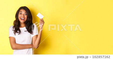 Portrait of amazed african-american girl, raising credit card and smiling excited, going on shopping, standing over yellow background 102857162