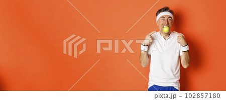 Portrait of funny, handsome middle-aged fitness guy, holding apple with teeth and pointing at it, eating healthy and working out, standing over orange background Portrait of funny, handsome middle-aged fitness guy, holding apple with teeth and pointing at it, eating healthy and working out, standing over orange background 102857180