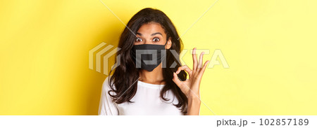 Concept of coronavirus, pandemic and lifestyle. Close-up of amazed and satisfied african-american girl, wearing black face mask, showing okay sign, like something good, yellow background 102857189