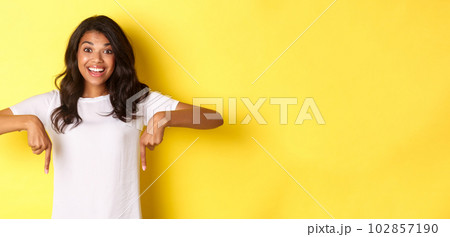 Portrait of happy african-american teenage girl showing banner, pointing fingers down at copy space and smiling excited, standing over yellow background 102857190