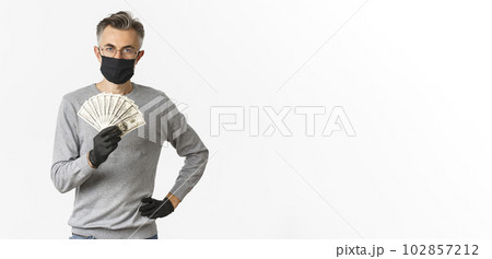 Concept of covid-19, social distancing and lifestyle. Portrait of successful middle-aged man in medical mask, gloves and glasses, showing money, standing over white background confident Concept of covid-19, social distancing and lifestyle. Portrait of successful middle-aged man in medical mask, gloves and glasses, showing money, standing over white background confident 102857212