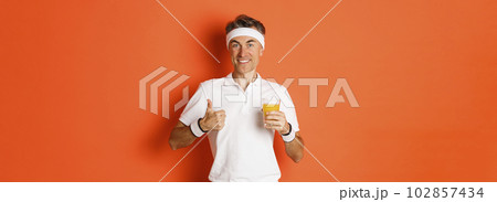 Concept of workout, gym and lifestyle. Image of fit middle-aged man, staying in shape with exercises and healthy food, holding fresh juice and showing thumbs-up, standing over orange background 102857434