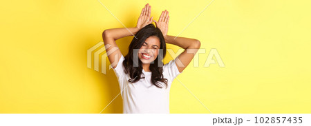 Portrait of attractive african-american girl, smiling and showing bunny ears on head, standing silly and coquettish over yellow background Portrait of attractive african-american girl, smiling and showing bunny ears on head, standing silly and coquettish over yellow background 102857435