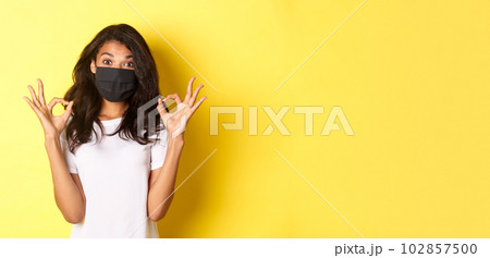 Concept of coronavirus, pandemic and lifestyle. Portrait of pleased african-american girl in black face mask, showing okay signs in approval, like something good, yellow background Concept of coronavirus, pandemic and lifestyle. Portrait of pleased african-american girl in black face mask, showing okay signs in approval, like something good, yellow background 102857500