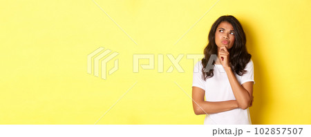 Portrait of attractive african-american woman thinking, making her choice, looking at upper left corner thoughtful and deciding, standing over yellow background Portrait of attractive african-american woman thinking, making her choice, looking at upper left corner thoughtful and deciding, standing over yellow background 102857507